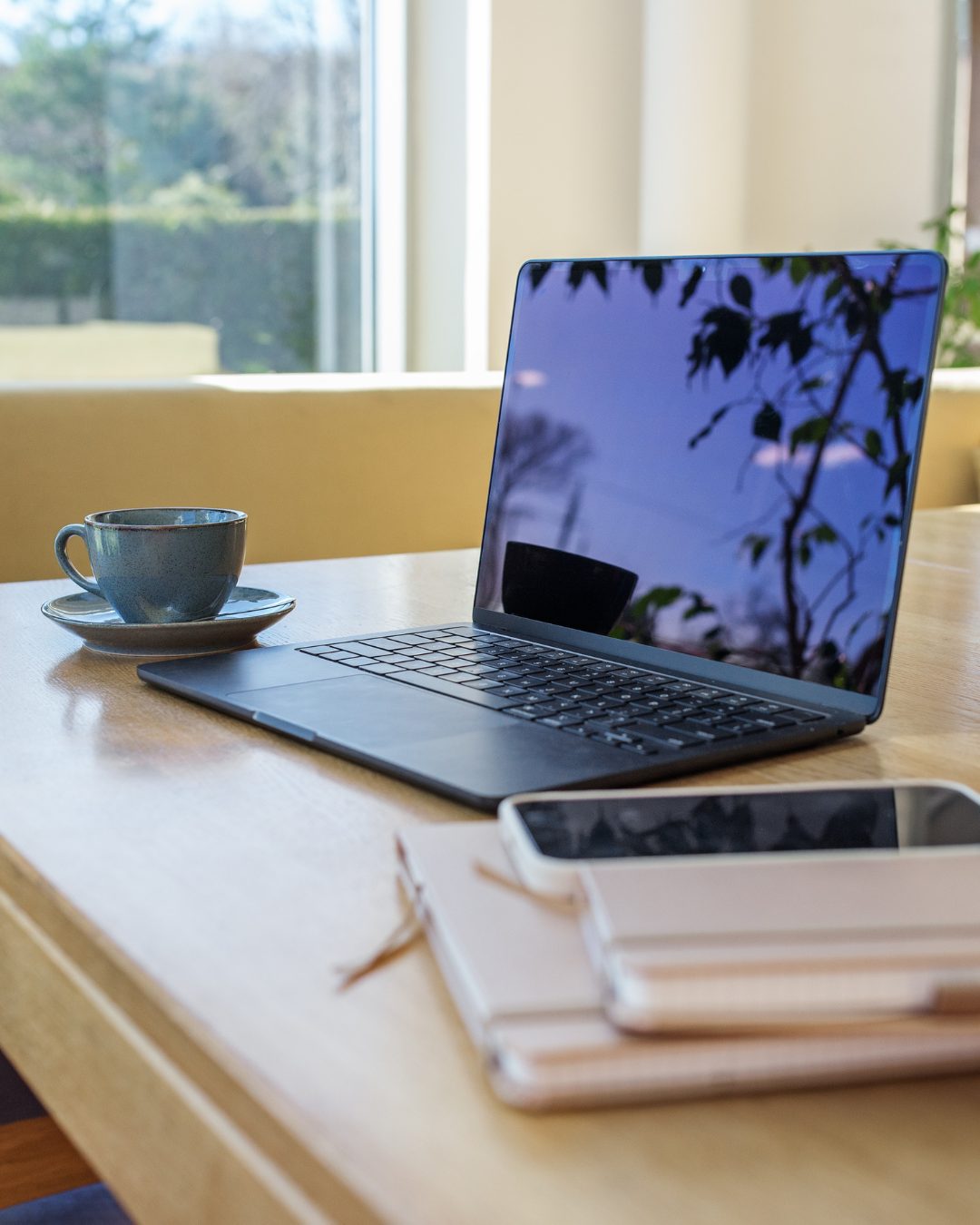 Laptop, coffee cup and notebooks on a wooden desk in a sunlit room
